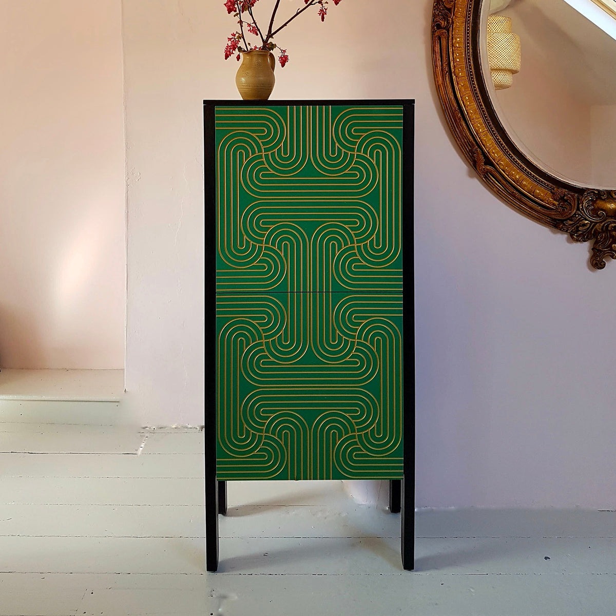 A tall green cabinet with a geometric wavy pattern, styled with a small vase of pink branches on top, set against a pale pink wall with a large ornate mirror on the right.