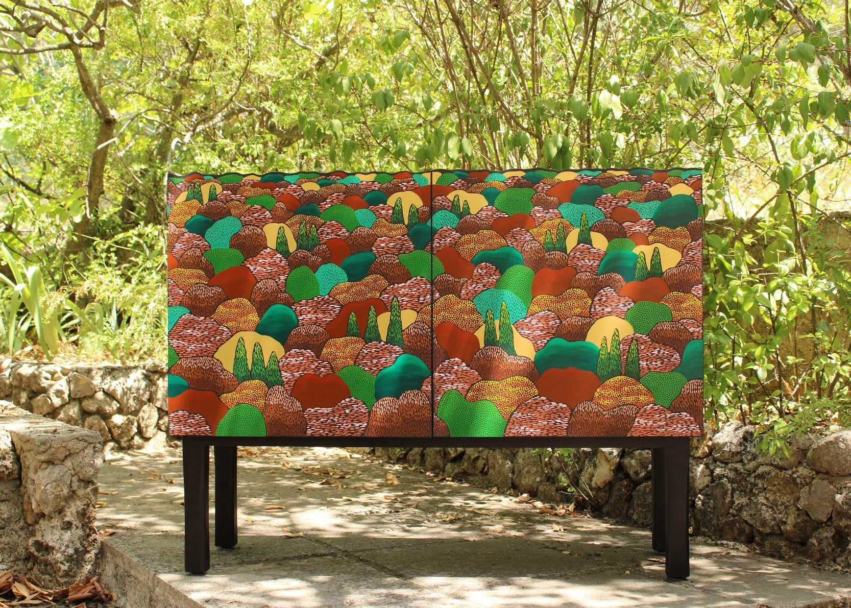 Colorful patterned cabinet in a natural setting with greenery and stone wall.