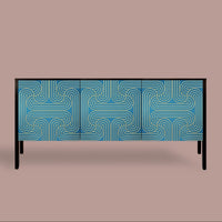 A modern three-door sideboard or credenza with a blue and gold geometric maze-like pattern on the doors, supported by slim black legs, set against a solid taupe background.