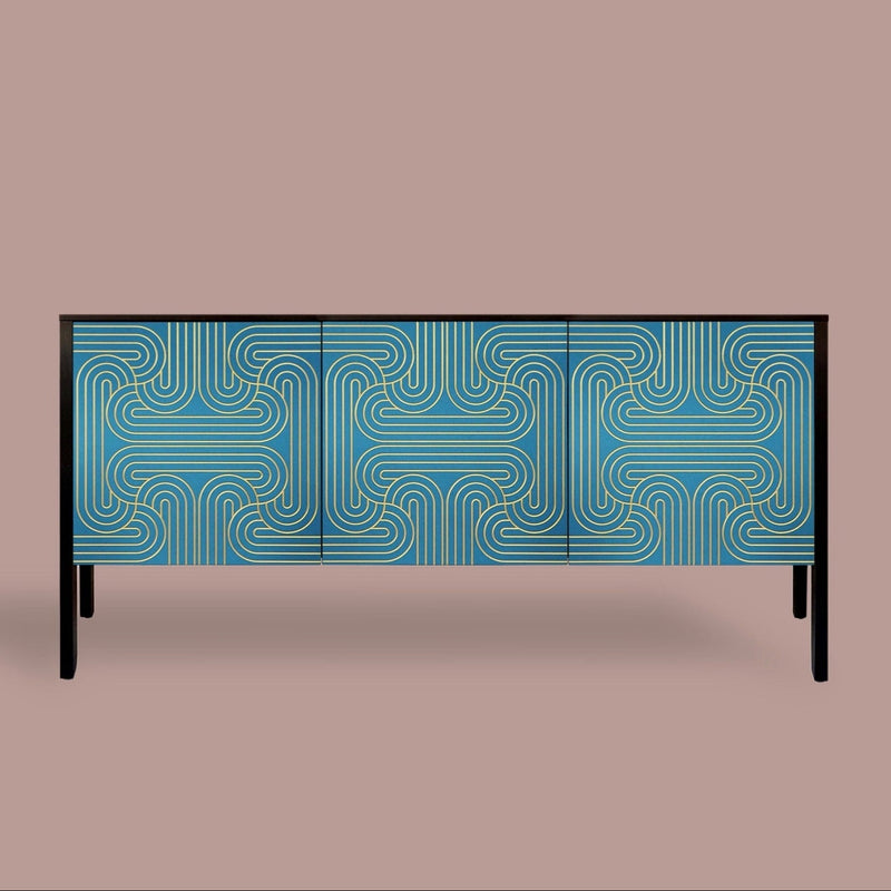 A modern three-door sideboard or credenza with a blue and gold geometric maze-like pattern on the doors, supported by slim black legs, set against a solid taupe background.