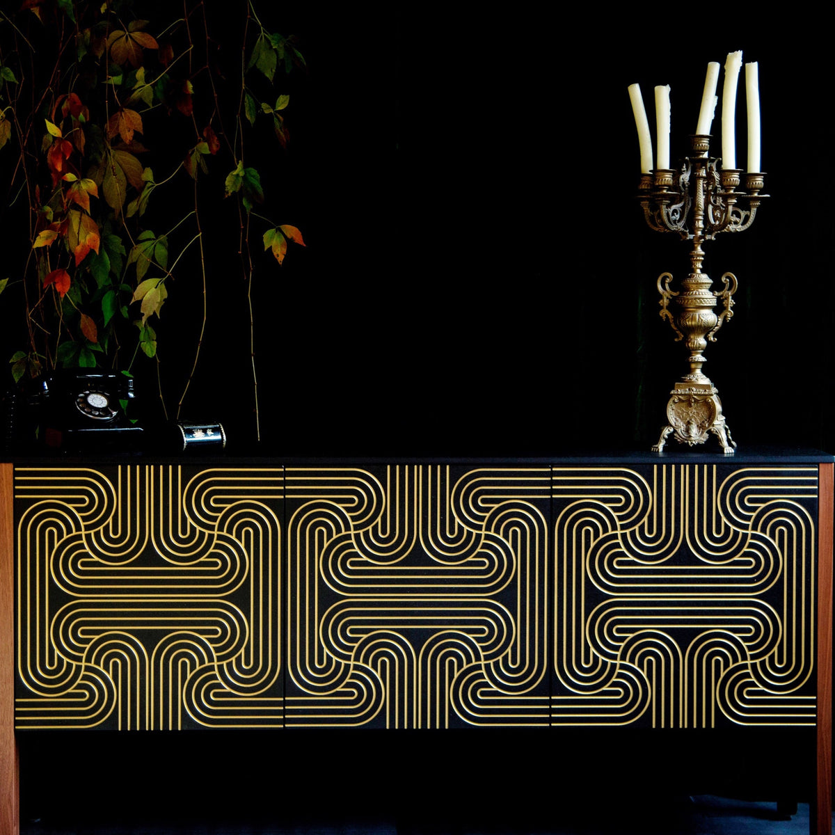 A black and gold sideboard with a bold geometric gold line pattern on the front, styled against a black background with a candelabra and hanging foliage.