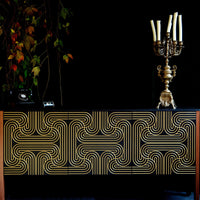 A black and gold sideboard with a bold geometric gold line pattern on the front, styled against a black background with a candelabra and hanging foliage.