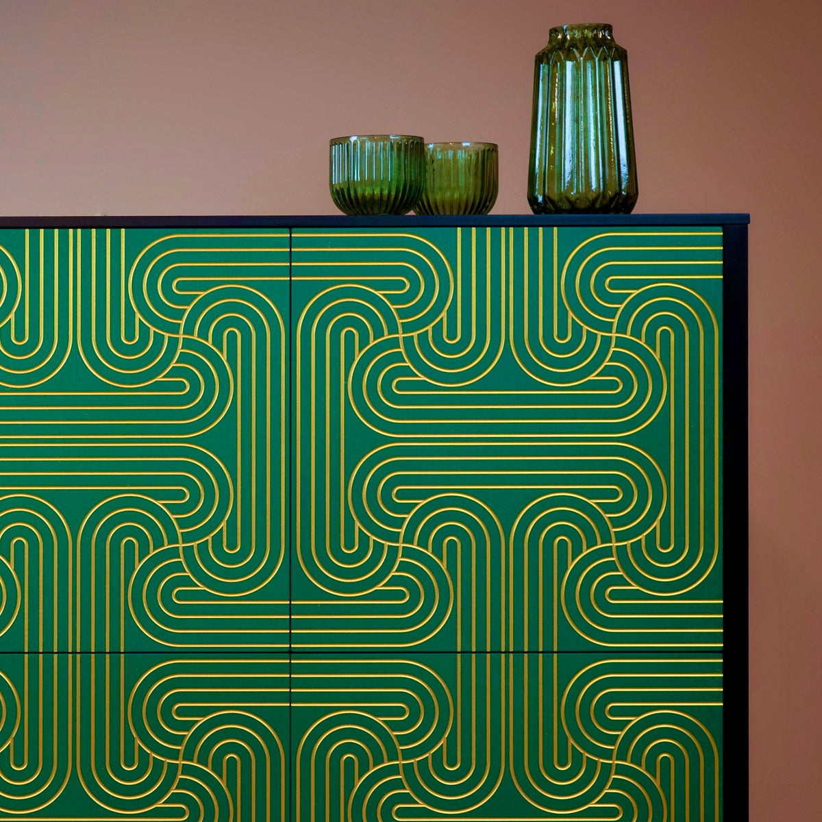 A modern sideboard or cabinet with green doors featuring a gold geometric maze-like pattern, topped with two small green ribbed bowls and a tall green glass vase against a solid warm brown background.