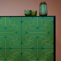 A modern sideboard or cabinet with green doors featuring a gold geometric maze-like pattern, topped with two small green ribbed bowls and a tall green glass vase against a solid warm brown background.