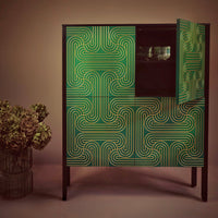 A modern bar cabinet with a dark wood frame and doors featuring a bold green and gold geometric maze-like pattern, shown next to a vase of dried flowers against a warm brown background.